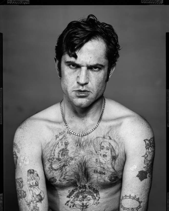 A man looks at the camera in a black and white photo. He is topless and has tattoos. He looks a little angry.